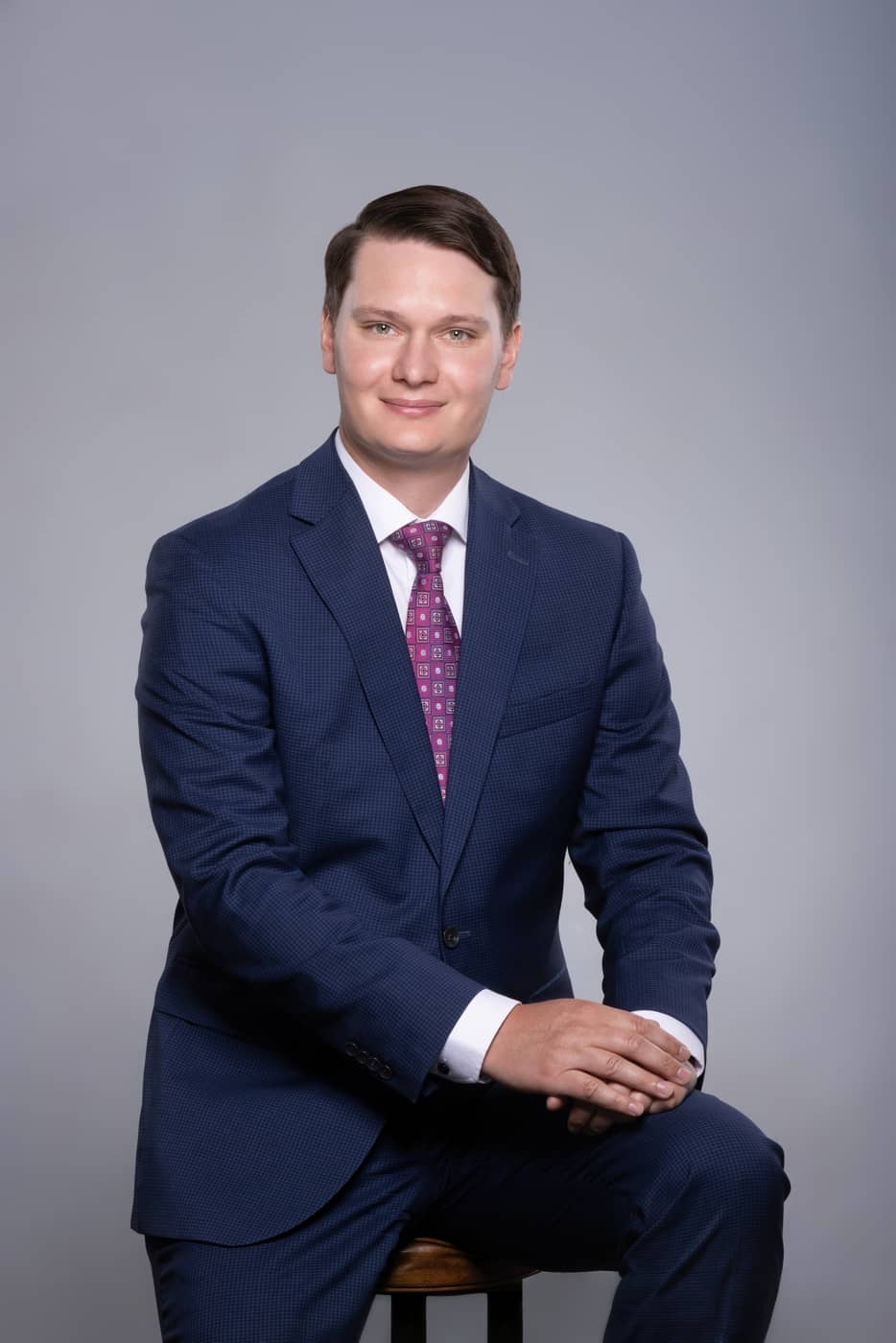 Seated portrait of Aaron in a navy suit with hands folded