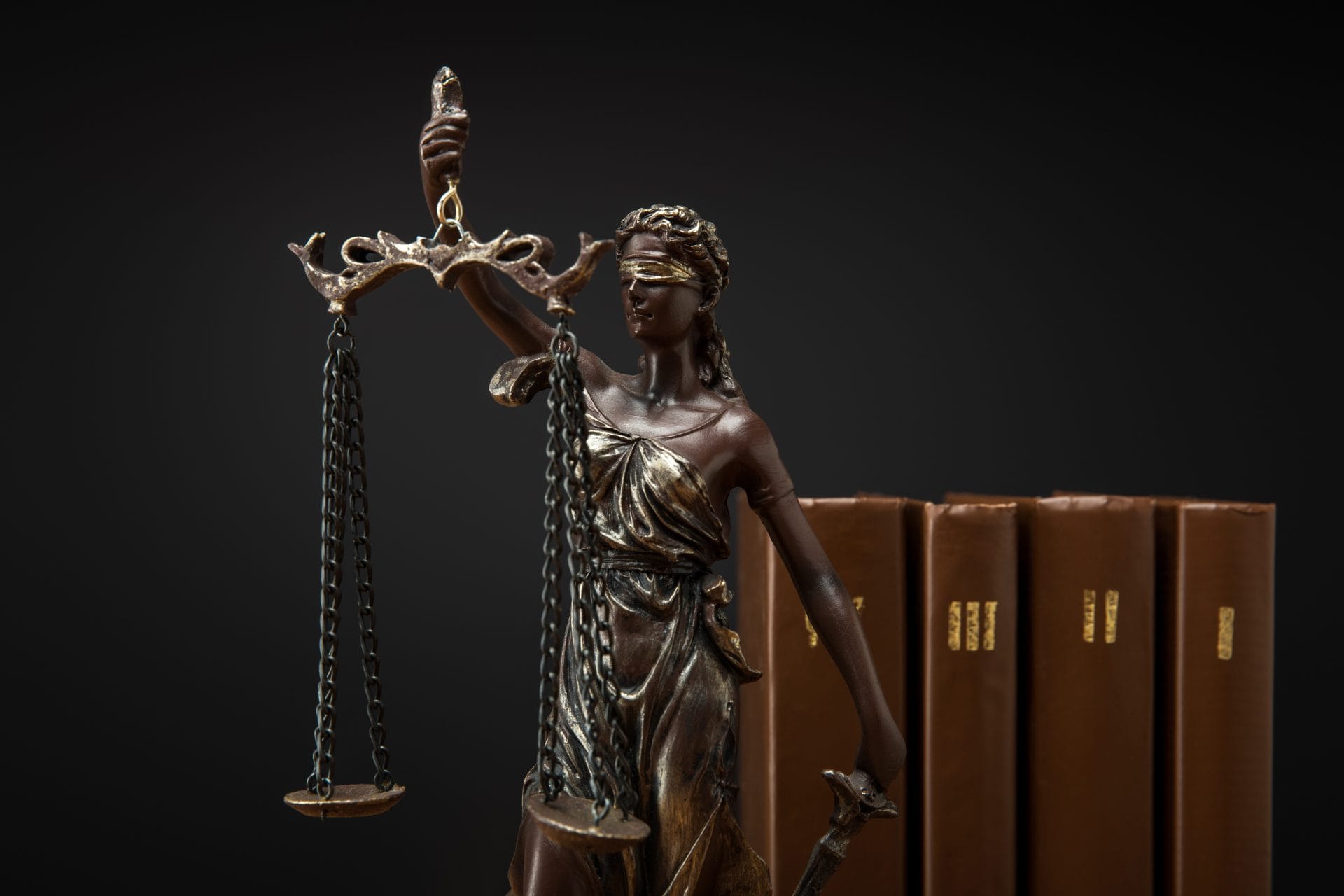 Blindfolded bronze Lady Justice statue holding scales beside a row of leather law books