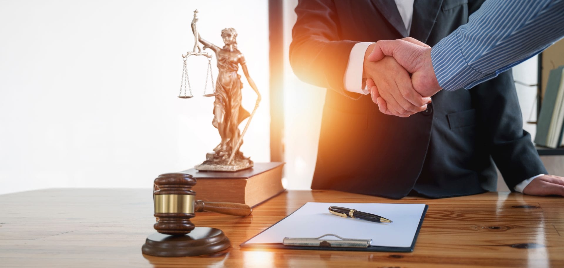 Two professionals shaking hands over a desk with a gavel, law book, and small justice statue