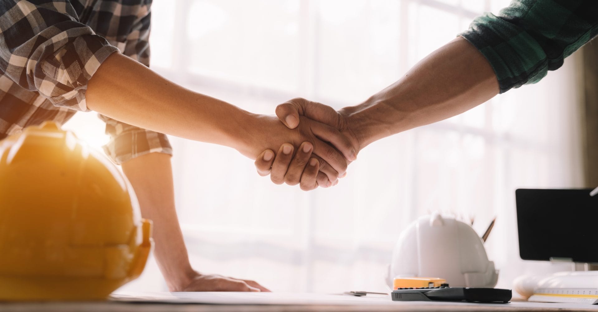 Two construction workers shaking hands with hard hats resting on the desk
